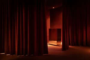Dark red velvet theater stage curtains in front of a dimly lit, minimalist interior with a mirrored box in the center.