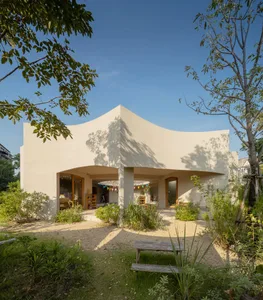 A modern house with a curved roof and concrete walls surrounded by greenery and a wooden bench in the foreground.