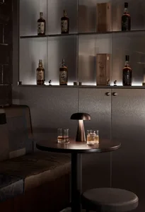 A bar counter with a metal finish, featuring a round table with two glasses of whiskey and a small lamp.