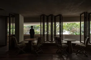 A coffee shop interior with wooden tables, concrete floors, and large glass windows overlooking greenery.