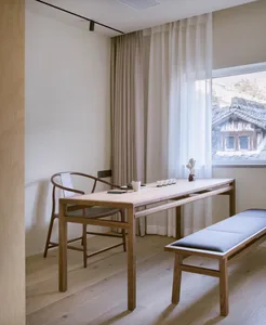 A minimalist interior featuring a wooden table and bench near a window with curtains and a wooden chair.