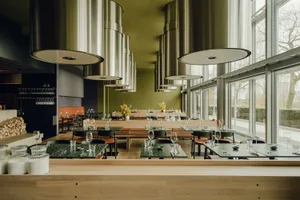 Modern industrial restaurant interior with metal light fixtures, oak benches, glass doors, and leather chairs.