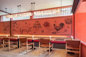 A modern restaurant interior featuring a vibrant red wall mural depicting various food items and the words "TE CHÓ VAYÓ."