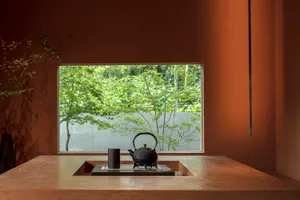 A minimalist interior featuring a teapot on a wooden table, with a window showing greenery outside.