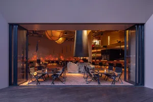 Industrially styled restaurant interior with wooden tables and chairs, a central fireplace, and warm lighting.