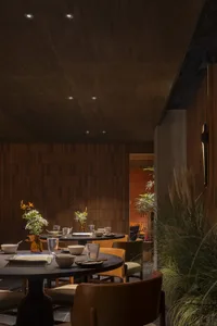 Modern restaurant interior with wooden tables and chairs adorned with plant arrangements, set with bowls, glasses, and books.