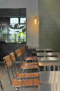 Industrial-style restaurant with metal and wooden chairs and tables, large windows, green plants, and warm lighting.