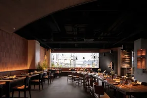 A modern restaurant interior with wooden tables, concrete walls, and industrial-style lighting.