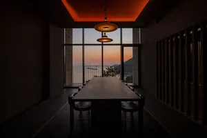 A modern dining room featuring a long wooden table and chairs, set against a large glass window overlooking a sunset over the