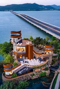 A castle-like building situated over a body of water, with a long bridge extending into the distance and mountains in the bac
