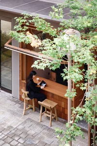 A person wearing black sits at a wooden table with a tablet and a cup of coffee in a modern outdoor coffee shop.