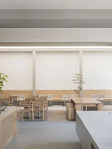 A minimalist restaurant interior featuring wooden tables and chairs with white accents, surrounded by a light-colored wooden