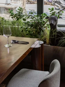 A modern restaurant interior featuring a wooden table with a wine glass, metal cutlery, and a glass window with green plants 