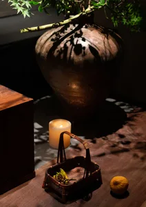A ceramic vase casting a shadow on a wooden table with a lit candle and basket of leaves.