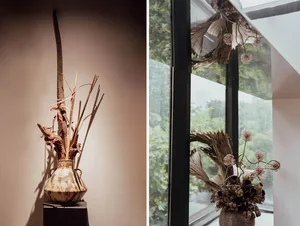 A vases adorned with dried plants sits on a wooden pedestal against a beige wall.
