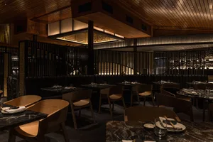 A modern restaurant interior featuring wooden tables and chairs, a wooden staircase, and a wooden ceiling.