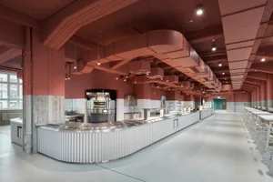 An industrial-style cafeteria with a long metal counter, white tile backsplash, and exposed ventilation systems.