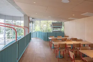 Industrial-themed restaurant interior with wooden tables and chairs, large windows and greenery outside.