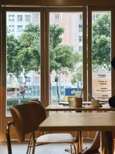 A cafe interior with modern wooden tables and chairs, featuring a window view of trees and buildings.