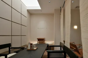 A minimalist restaurant interior featuring a wooden dining table and chairs, soft lighting, and a skylight.