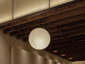 A hanging spherical paper lantern illuminated from within, hanging from wooden beams in a minimalist interior setting.