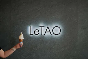 A hand holding an ice cream cone in front of a gray marble sign with 'LeTAO' illuminated in the background.