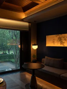 A modern living room with wooden furniture, fabric seating, and glass elements, featuring trees outside a large window.