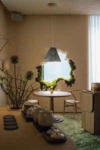 A modern restaurant interior with natural lighting, wooden tables, chairs, and a hanging pendant light surrounded by green mo