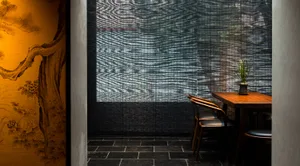 A minimalist restaurant interior featuring a wooden table, black cushions, and a bamboo shade covering a large window.