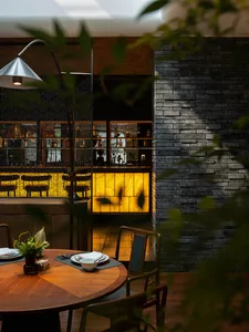Modern industrial-style restaurant interior with a brick wall backdrop, round wooden dining table, and a bar or lounge area i