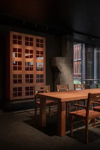 A minimalist office space featuring a wooden table and glass cabinet, with a dark carpet and concrete walls.