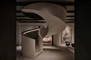 A minimalist bedroom connected by a spiral staircase made of concrete, featuring a bed with a beige headboard and a large abs