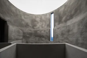An image depicting a minimalist concrete interior design with a large circular skylight and a tall, narrow window.