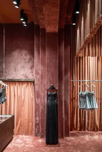 A modern retail storefront with velvet walls and curtains, featuring a mannequin wearing a black dress and hanging clothing i