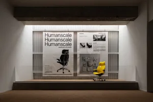 A Humanscale product display featuring a yellow office chair on a step platform with informational posters behind it.