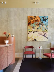 A modern restaurant lobby features a concrete reception desk with pink tiles and red cushioned chairs, alongside an abstract 