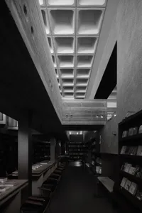 A black and white photograph of a modern library interior with concrete walls and a grid ceiling.