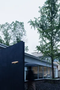A modern social club with a black wall, glass windows, and steel accents, surrounded by green trees and gravel.