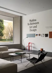 Modern minimalist living room with concrete, leather, and metal furniture, large window, and wall text reading 'Mutina In-Bet