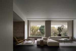 A modern minimalist living room featuring a large sectional sofa, a wooden chair, a coffee table, and large windows overlooki