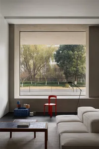 A modern living room featuring concrete walls and floor, with a red chair, white sofa, and a large window overlooking a green