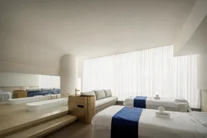 A serene spa room with white cloth textiles on stone flooring, glass walls, and a large window.