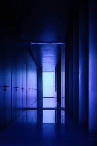 A long, narrow corridor with walls illuminated in a single, deep blue hue, extending towards a bright endpoint.