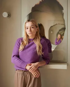 A woman with blonde hair wearing a purple sweater and gold jewelry standing in front of a mirror with a surreal reflection.