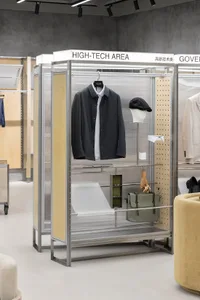 A high-tech clothing shop interior displaying a black jacket, cap, and green bag on a metallic rack with wooden backing.