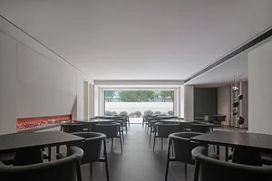 A sleek, minimalist restaurant interior featuring modern concrete dining tables and chairs arranged in rows, with a large flo