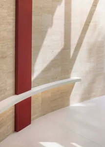 A minimalist interior with a red column and white bench, featuring natural light and shadows on a travertine wall.