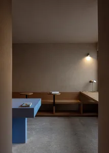 A minimalist workspace featuring a blue desk on a concrete floor with a wooden bench and built-in shelves.