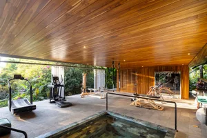 A modern home gym with wooden flooring and ceiling, featuring exercise equipment and a pool area.
