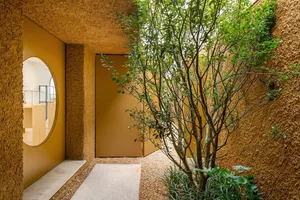 A minimalist entrance featuring textured concrete walls, a round window, a tree, and a gravel path.
